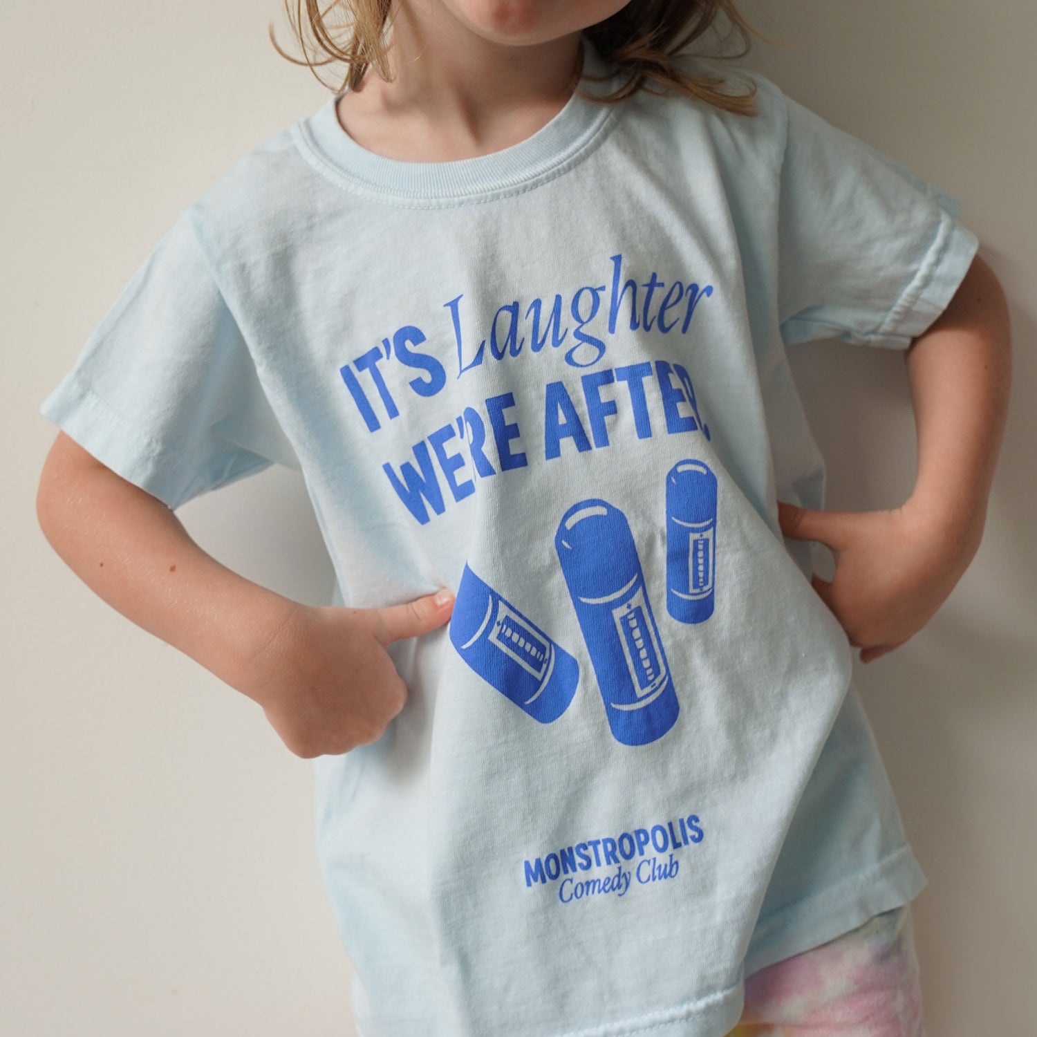 Front view of kids’ Disney-inspired Monstropolis Comedy Club t-shirt featuring the phrase ‘It’s Laughter We’re After’ above three laugh canisters with Monstropolis Comedy Club text underneath.