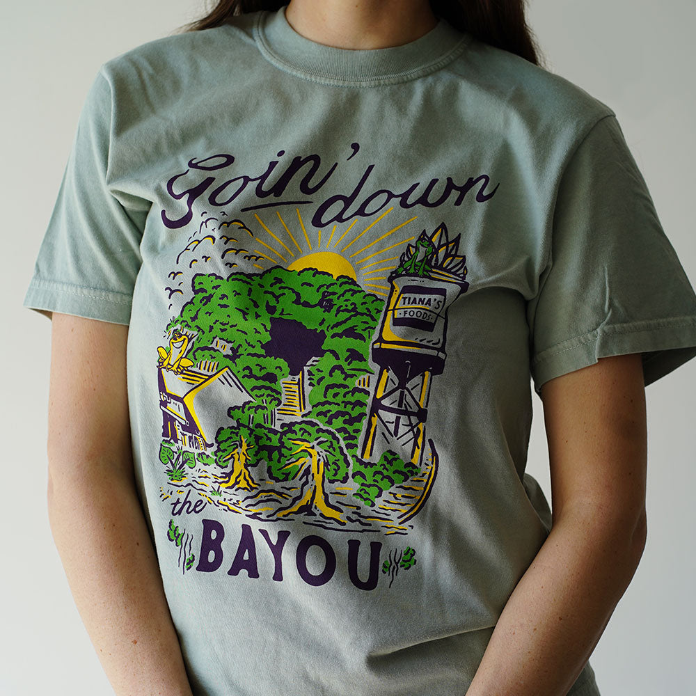 Front view of Disney-inspired Goin’ Down the Bayou t-shirt featuring text ‘Goin’ Down the Bayou’ above artwork of Tiana’s Bayou Adventure ride exterior with the big drop, Tiana’s Foods water tower, and cypress trees.