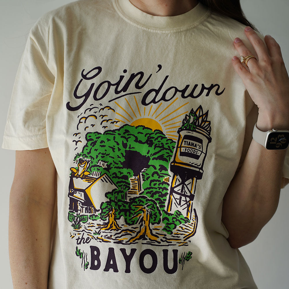 Front view of Disney-inspired Goin’ Down the Bayou t-shirt featuring text ‘Goin’ Down the Bayou’ above artwork of Tiana’s Bayou Adventure ride exterior with the big drop, Tiana’s Foods water tower, and cypress trees.