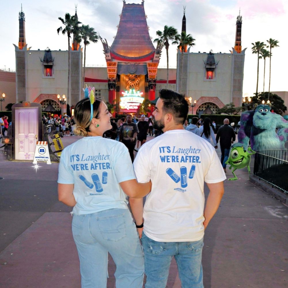 Couple at a Disney park wearing Monstropolis Comedy Club t-shirts. Woman in blue version and man in ivory version show the back design with the phrase ‘It’s Laughter We’re After,’ three laugh canisters, and Monstropolis Comedy Club text beneath.